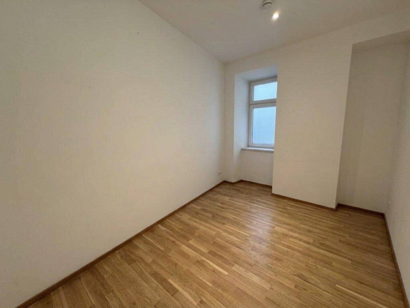 Kleiner, heller Raum mit Holzboden und einem einzelnen Fenster, ideal als Schlaf- oder Arbeitszimmer.