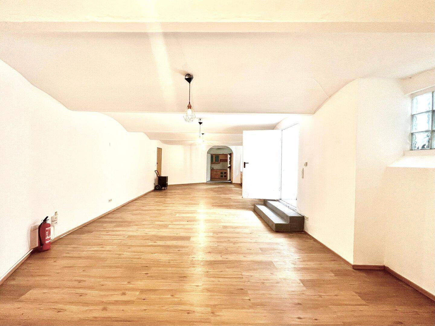 Weitläufiger Mehrzweckraum mit Holzboden, weiß gestrichenen Wänden und Zugang zum Außenbereich.