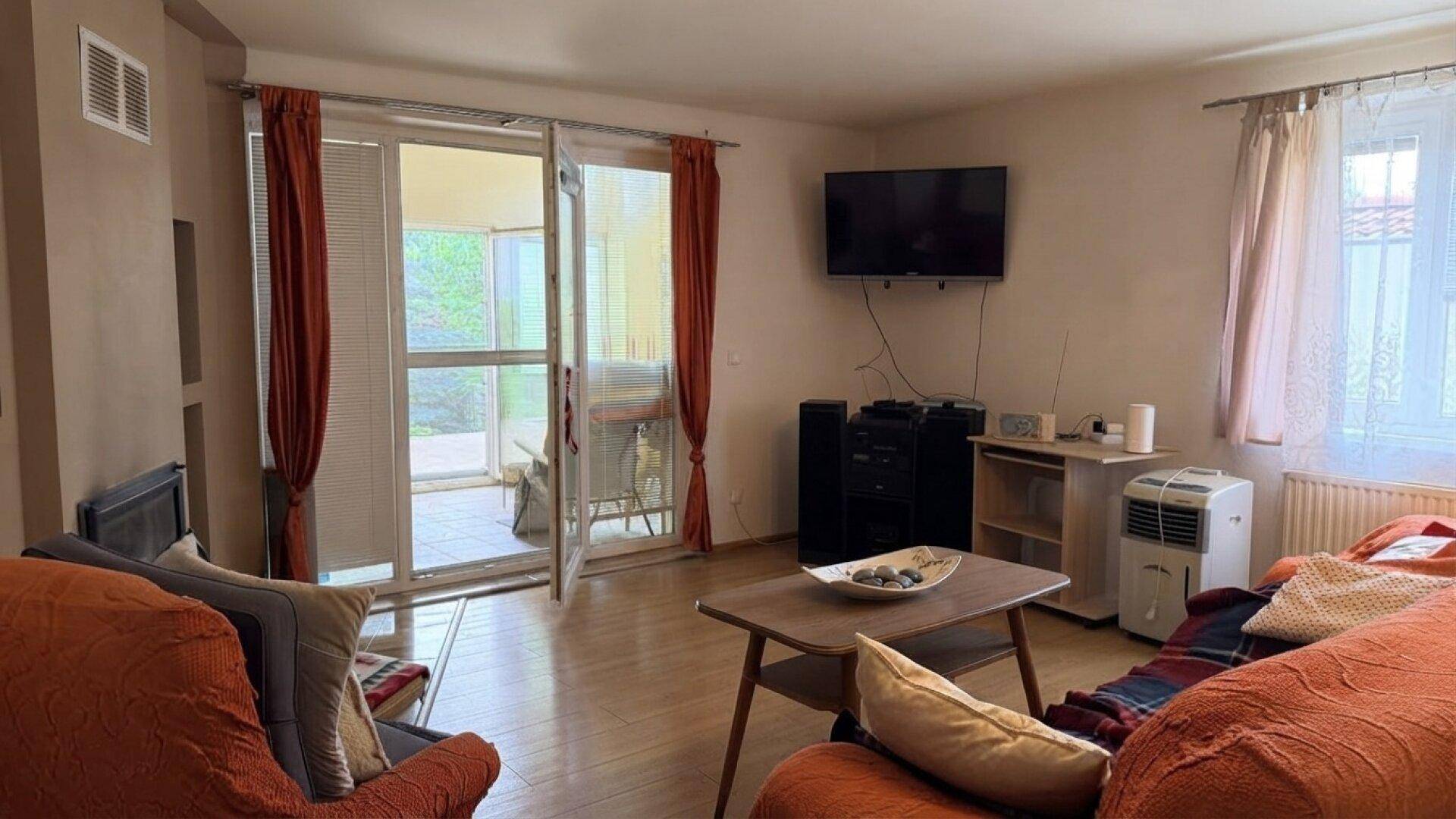 Wohnzimmer mit Holzboden, Fernseher und großen Schiebetüren, die zu einem Außenbereich führen.
