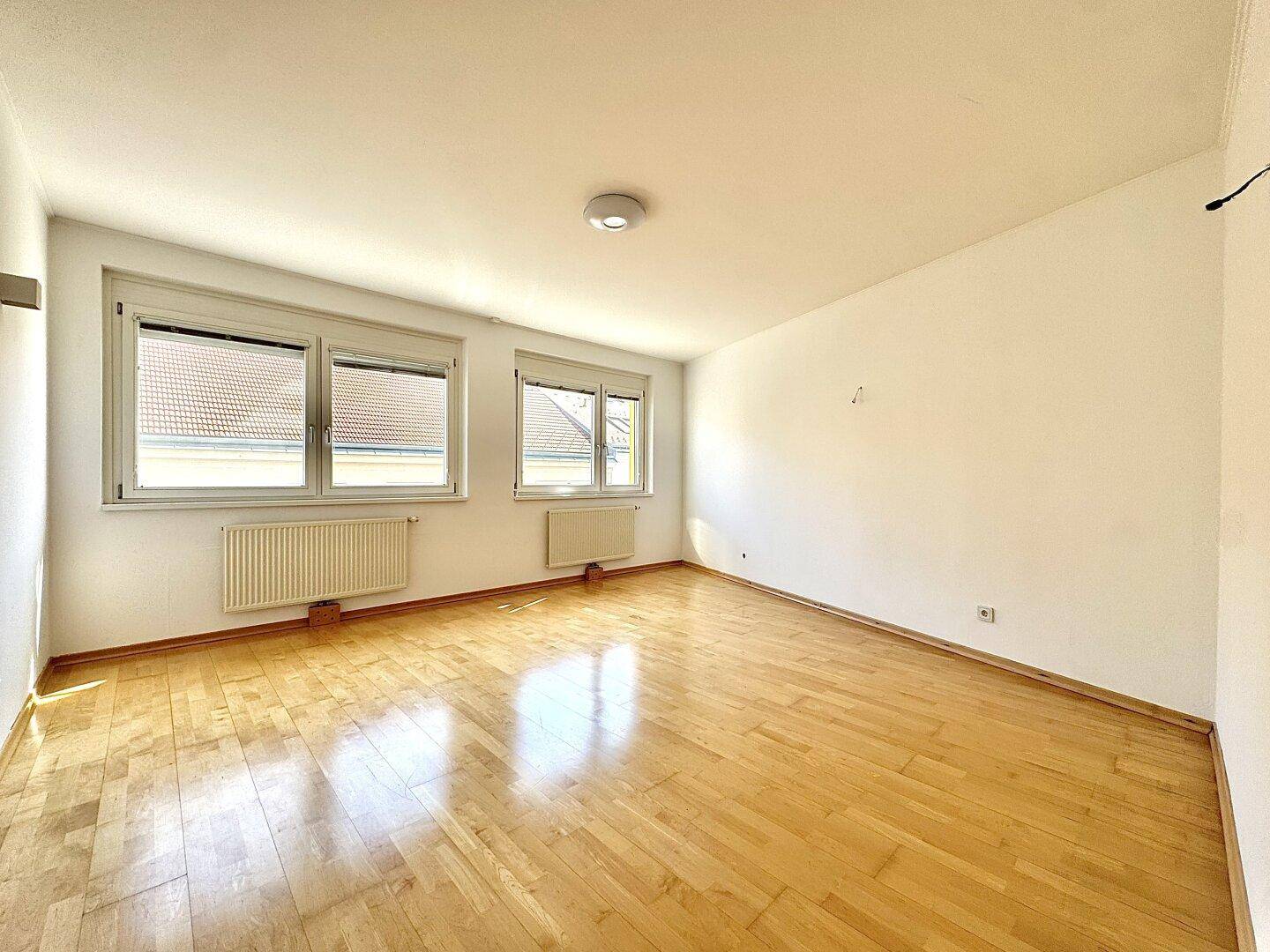 Helles Zimmer mit Holzfußboden, weißen Wänden und Blick ins Grüne durch die Fenster.