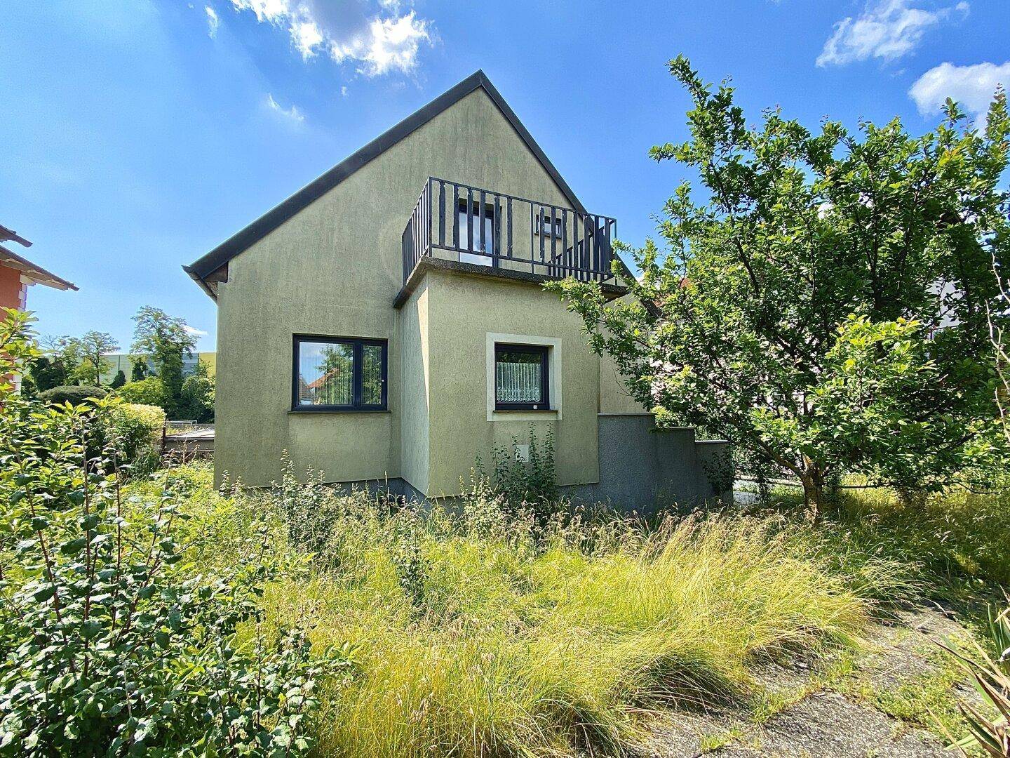 Rückansicht des Hauses mit Balkon und einem verwilderten Gartenbereich unter blauem Himmel.