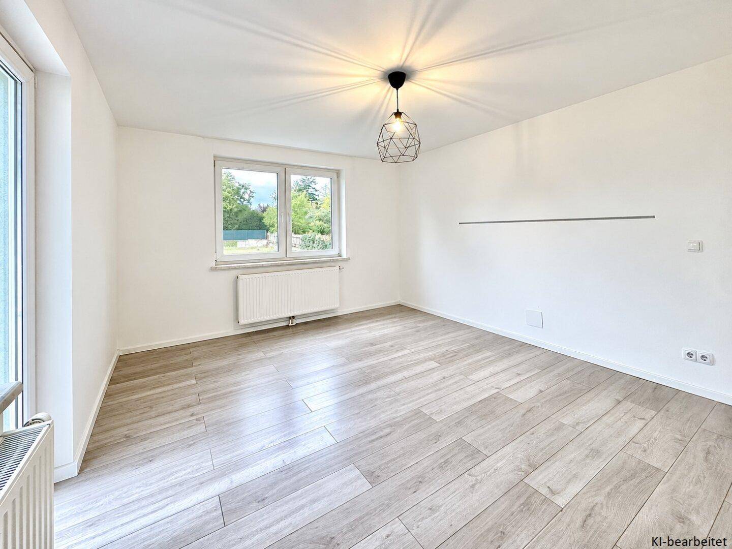 Helles, leeres Zimmer mit großem Fenster, hellem Laminatboden und moderner Deckenleuchte.