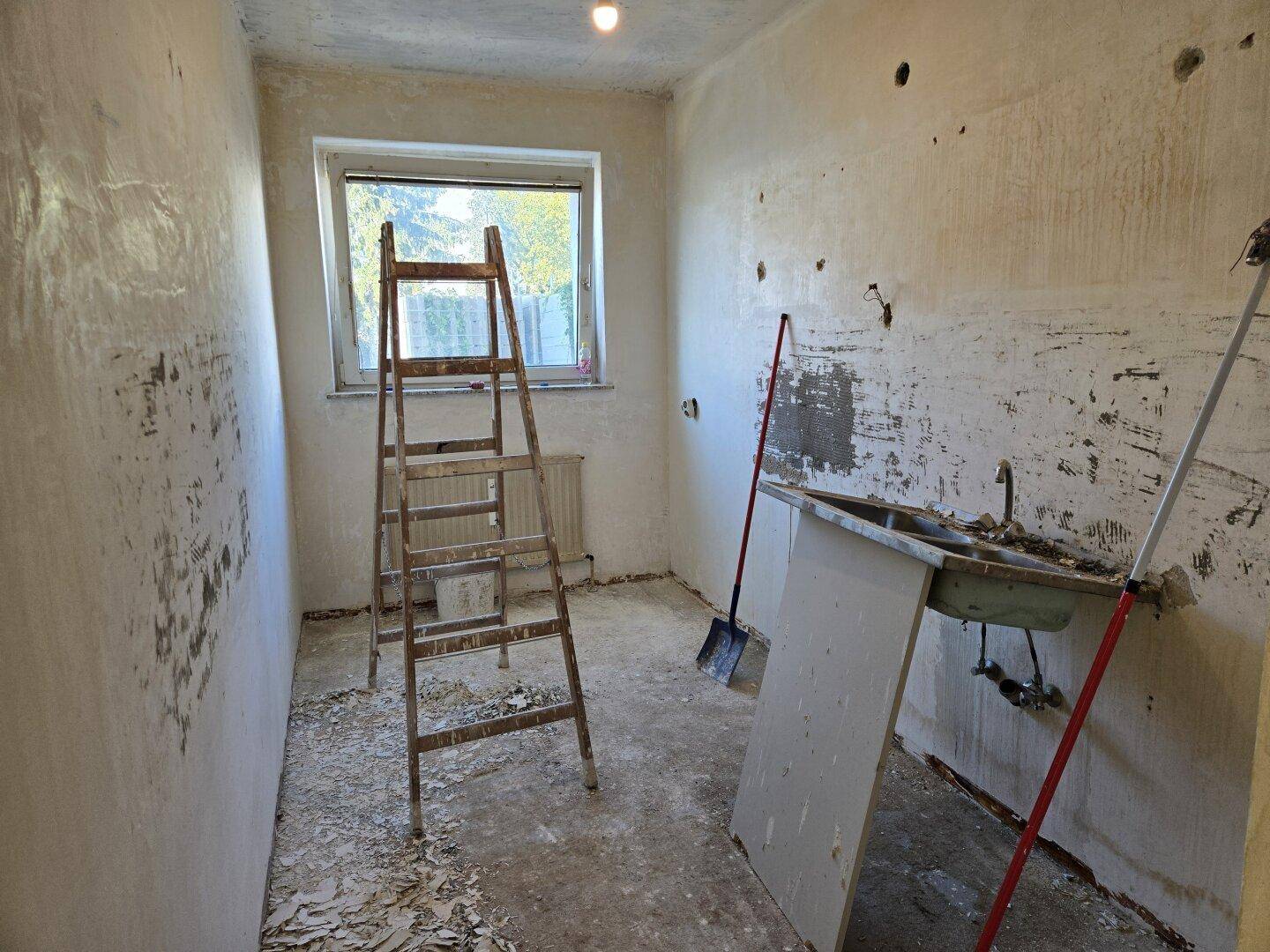 Küche im Rohbauzustand mit altem Spülbecken, Leiter und Fenster, bereit für die Renovierung.