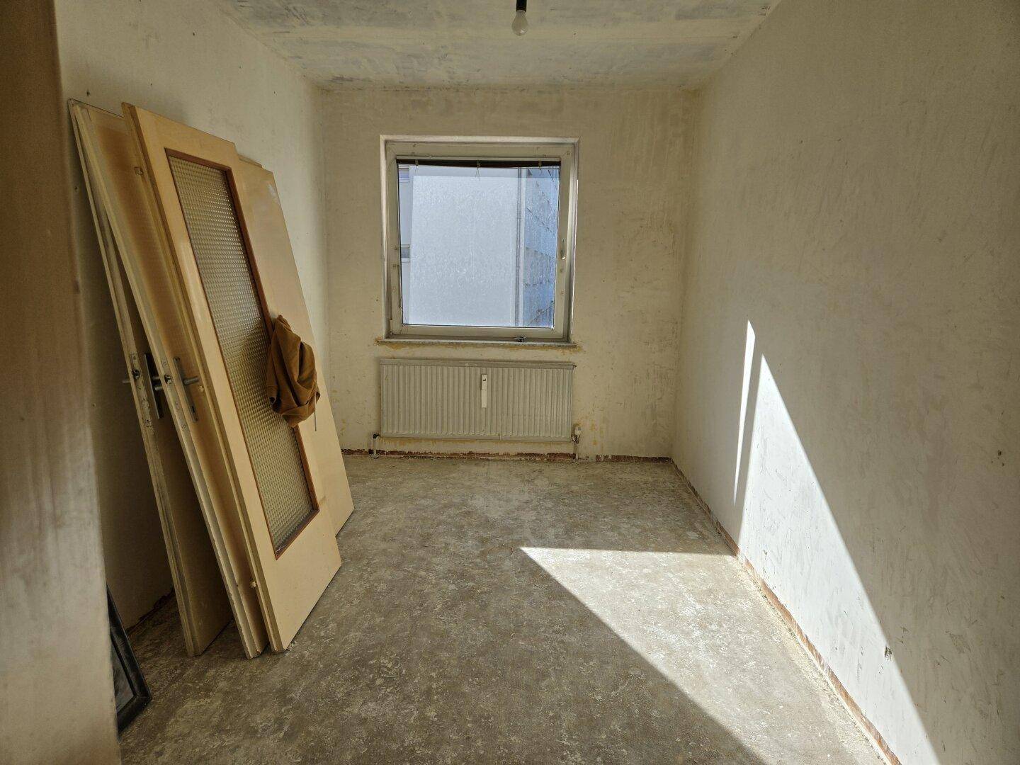 Kleiner Raum im Rohbau mit Fenster, Heizkörper und alten Türen, die auf die Installation warten.