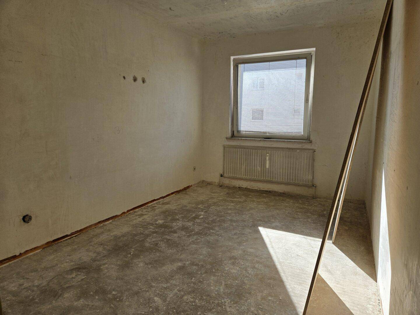Zimmer im Rohbauzustand mit Fenster, Heizkörper und viel natürlichem Licht.