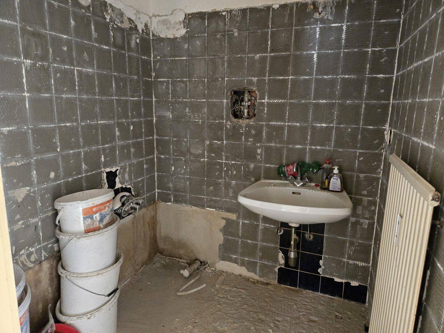 Badezimmer im Rohbau mit alten Fliesen, Waschbecken und Heizkörper, wartet auf Modernisierung.
