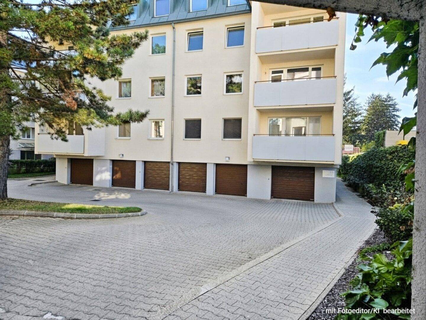 Mehrfamilienhaus mit Garagen im Erdgeschoss, Balkonen und einem gepflasterten Parkplatzbereich.