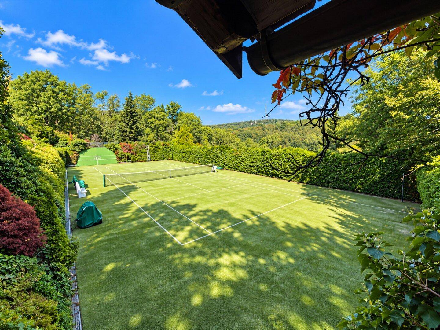 Blick auf den Tennisplatz von einer erhöhten Position, umgeben von hohen Hecken und grünen Hügeln.