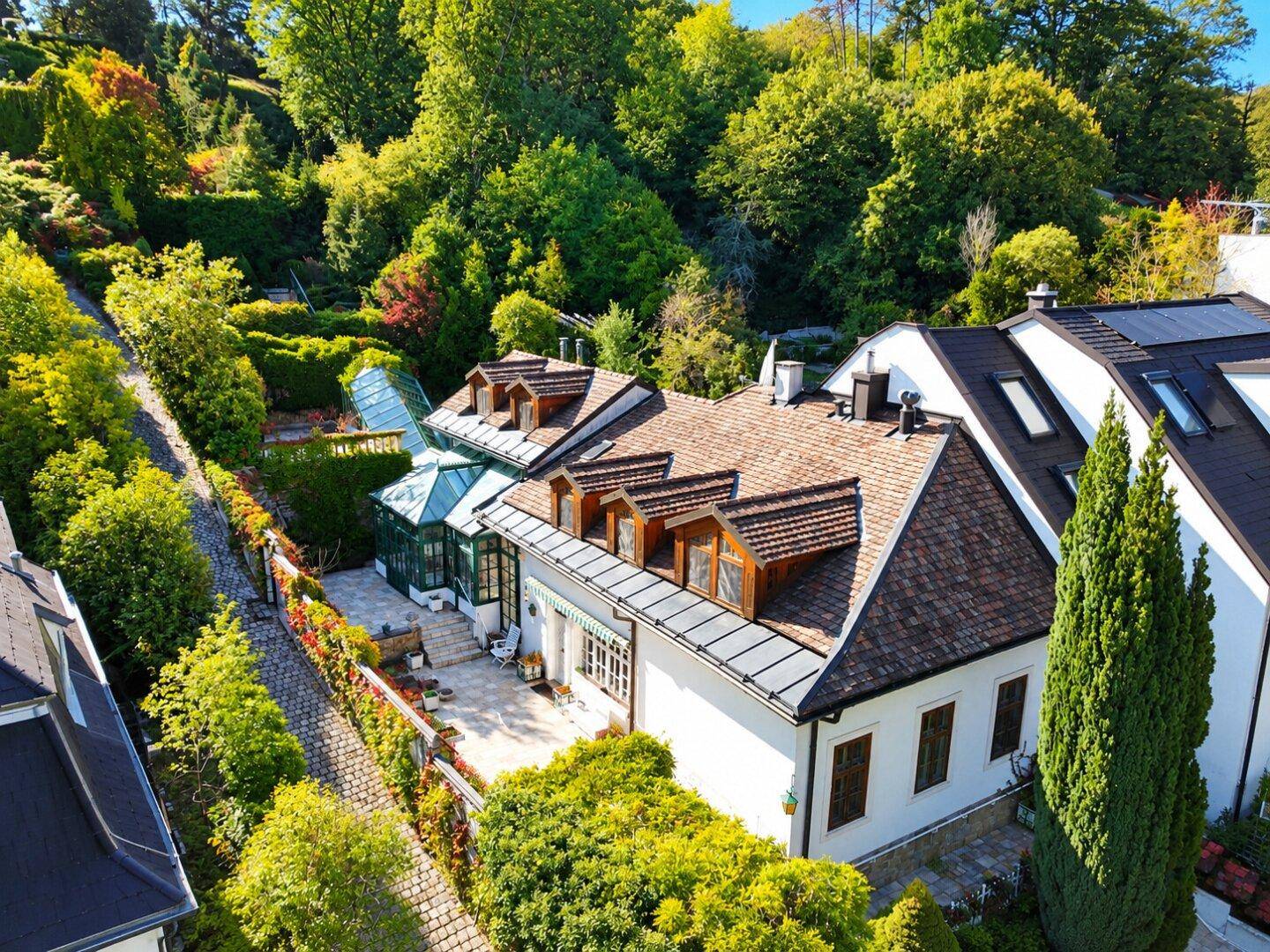 Luftaufnahme eines Hauses mit Mansarddach, großem Wintergarten und gepflasterter Terrasse, umgeben von viel Grün.