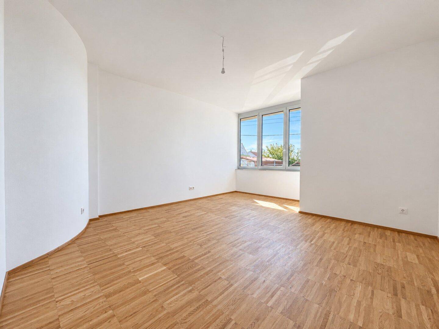 Helles, leeres Schlafzimmer mit Parkettboden und großen Fenstern, die viel Tageslicht hereinlassen.