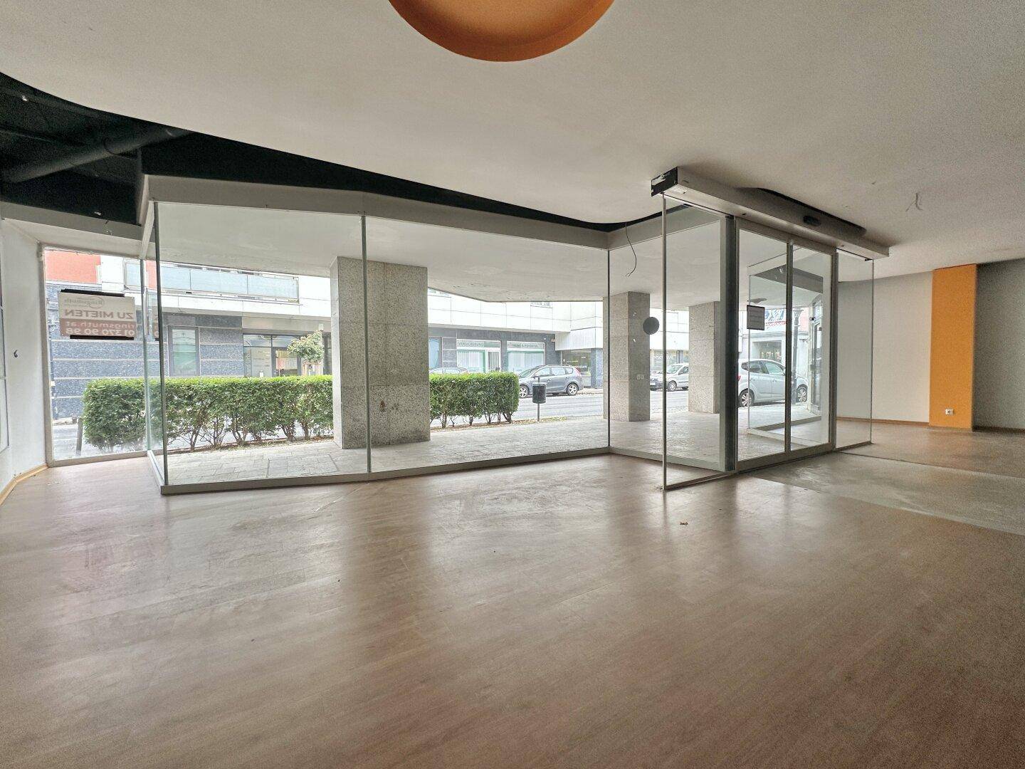 Leerer Gewerberaum mit großer Glasfront zur Straße und hellem Boden in Holzoptik.