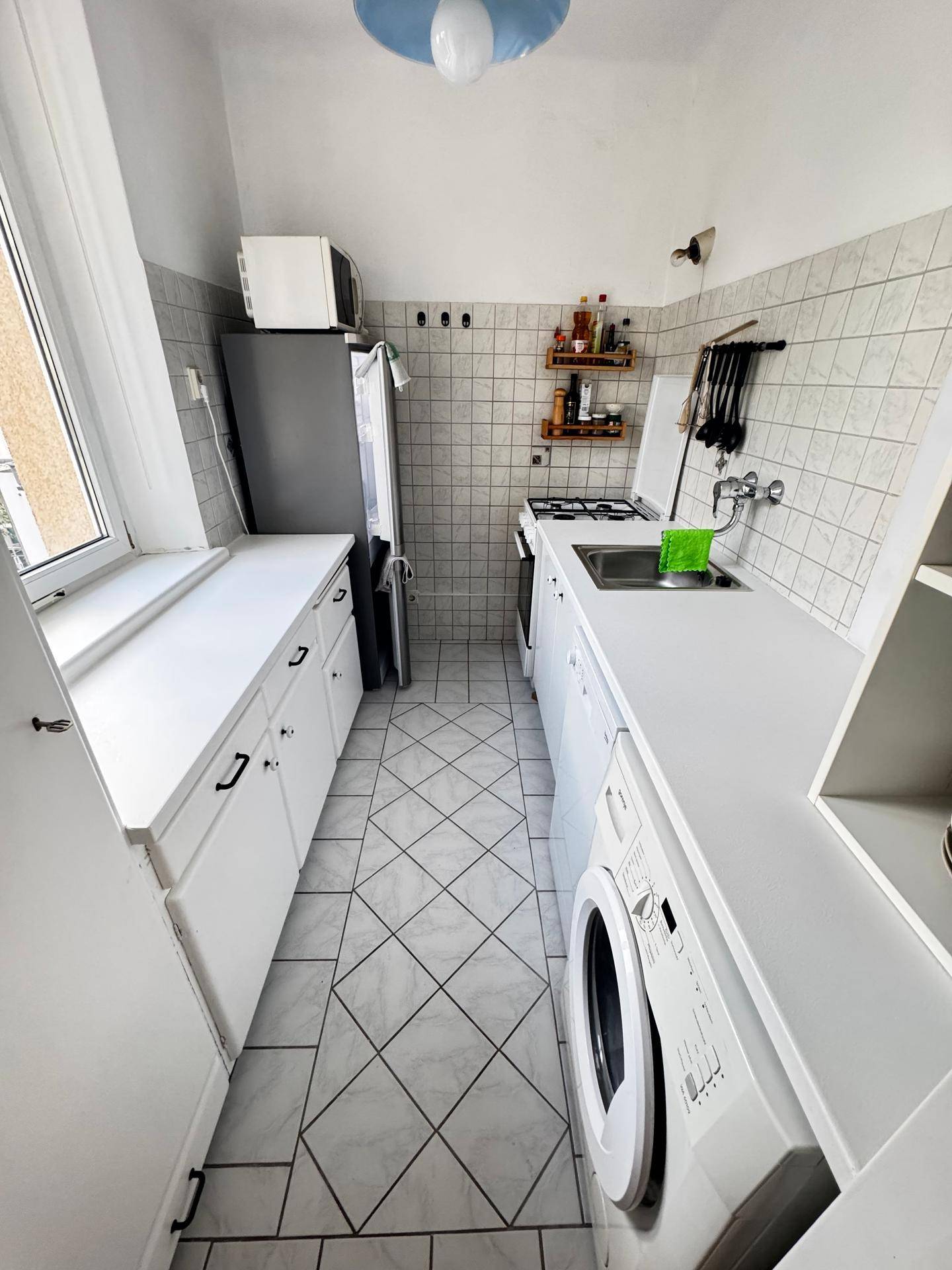 Schmale Küche mit weißen Schränken, Fliesenboden, Fenster und integrierten Geräten.