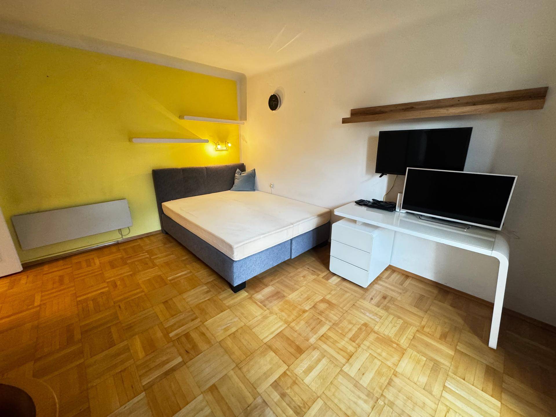 Schlafbereich mit Bett, Schreibtisch und gelber Akzentwand in einem hellen Raum.