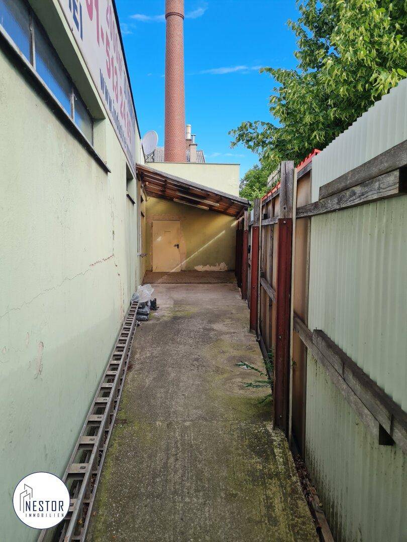 Schmaler Außengang oder Gasse zwischen Gebäuden und Zäunen mit einem kleinen überdachten Bereich am Ende.