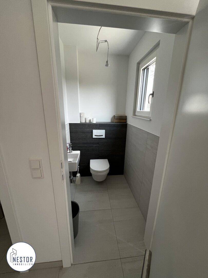 Modernes Gäste-WC mit Wand-WC, kleinem Waschbecken und Fenster für natürliche Belüftung.