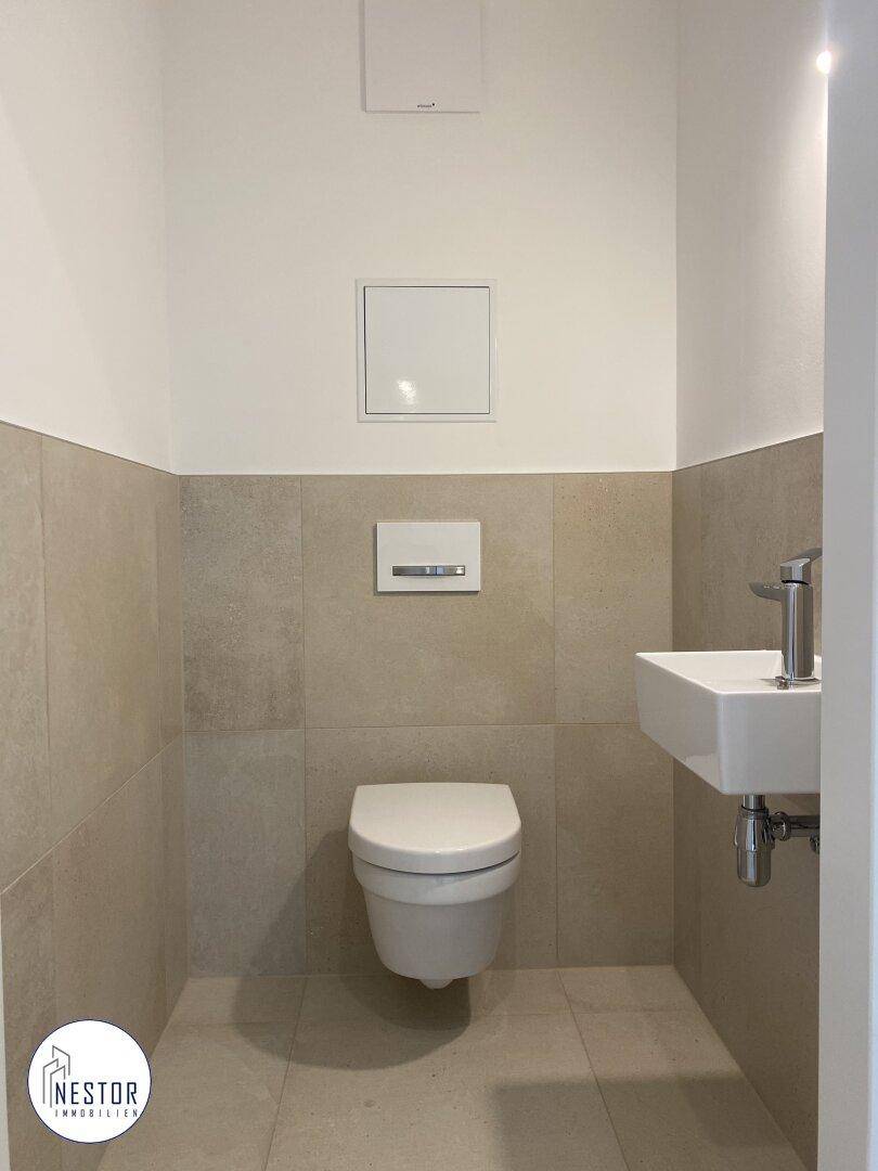 Modernes Gäste-WC mit beigen Fliesen, wandhängendem WC und kleinem Waschbecken.