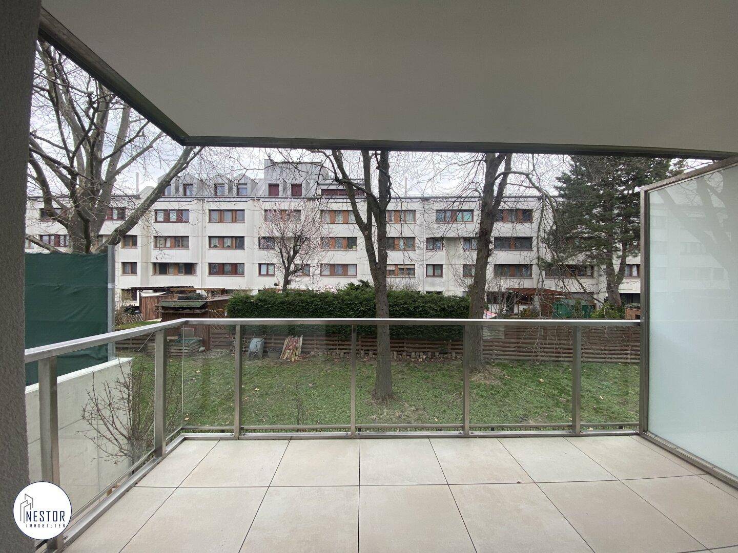 Moderner Balkon mit Fliesenboden und Glasgeländer, bietet Blick auf eine Grünfläche und umliegende Gebäude.