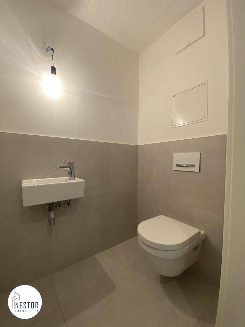Modernes Gäste-WC mit wandhängender Toilette, kleinem Waschbecken und grauen Fliesen.