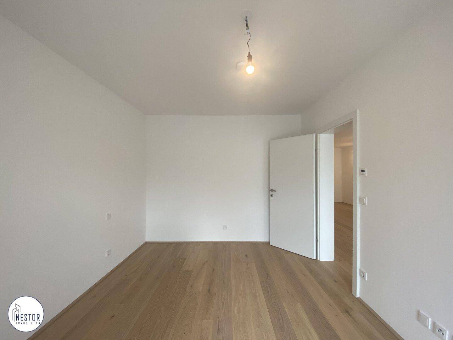 Leeres Schlafzimmer mit hellem Holzboden und weiß gestrichenen Wänden, mit offener Tür.