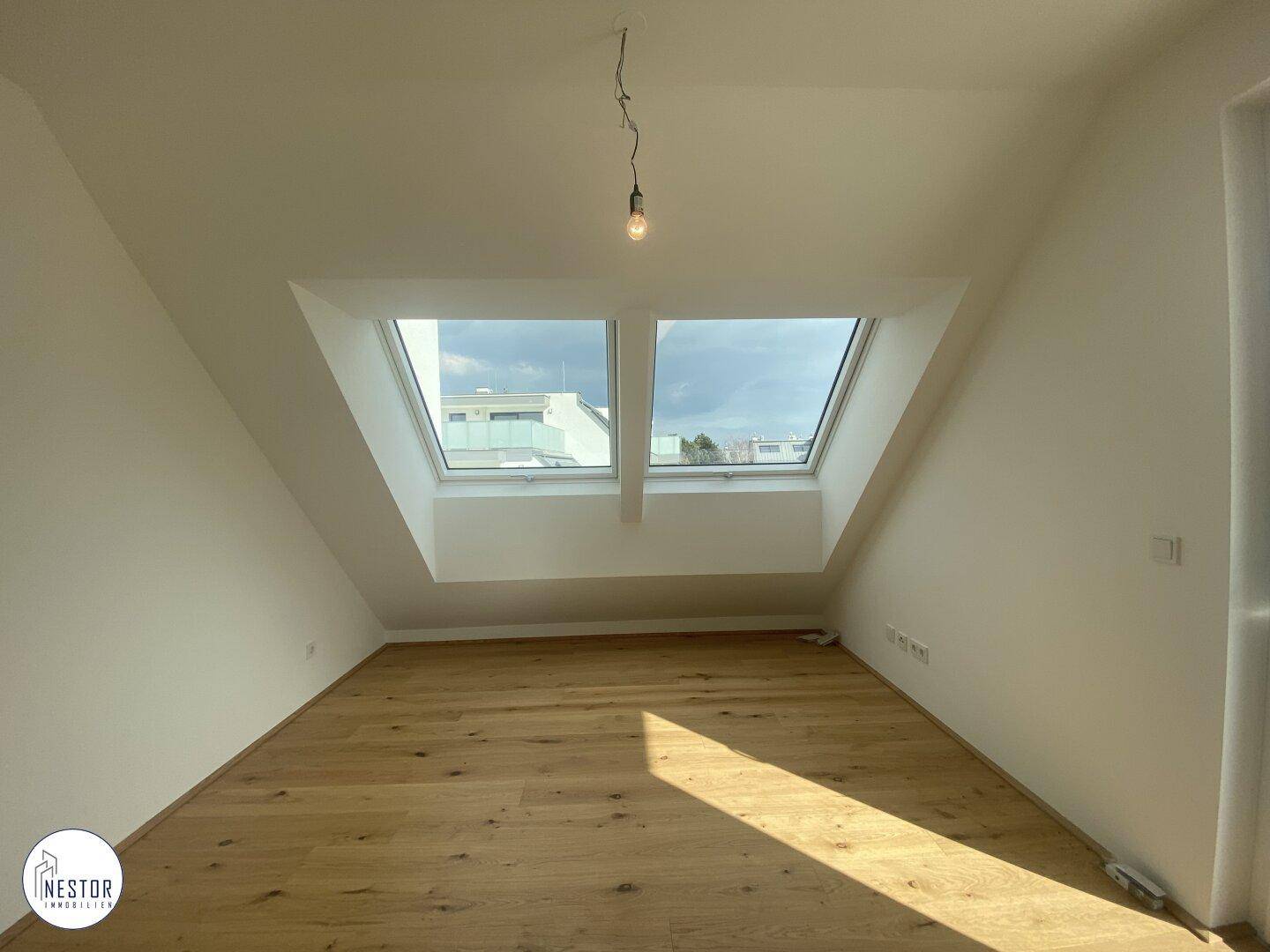 Leerer Raum im Dachgeschoss mit Holzboden und zwei großen Dachfenstern, die viel Licht hereinlassen.