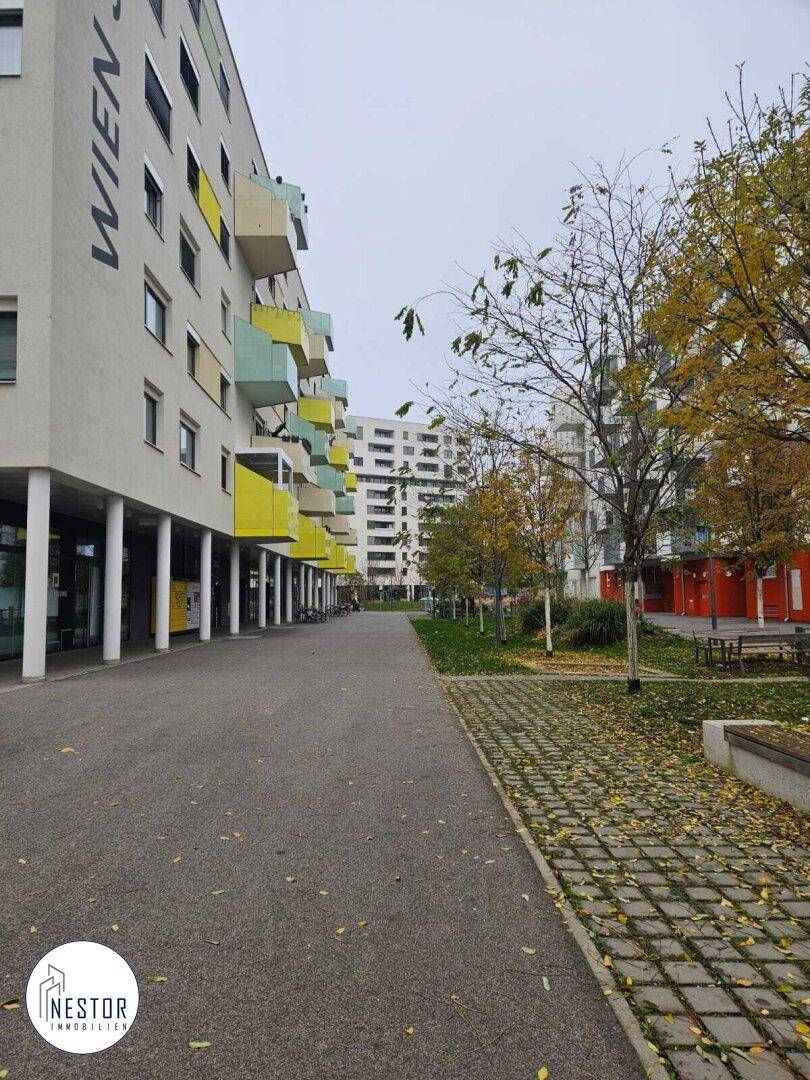 Seitenansicht eines modernen Wohngebäudes mit farbenfrohen Balkonen und einer breiten Fußgängerzone. Die Umgebung ist grün und einladend.