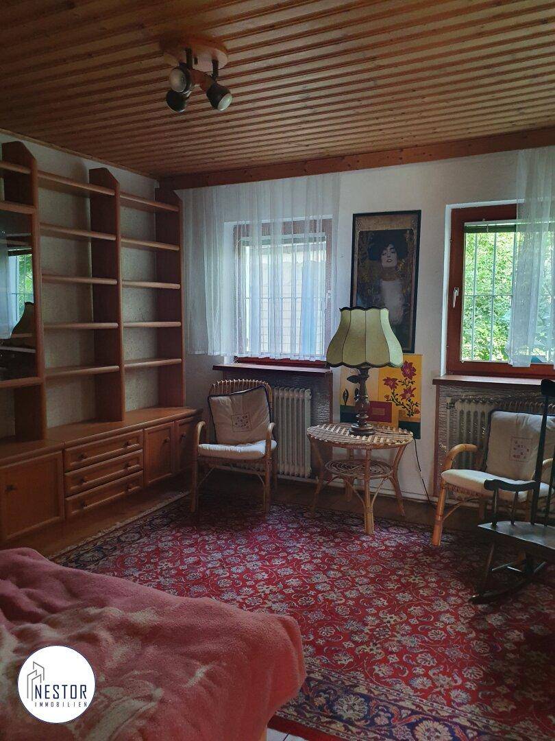 Gemütliches Zimmer mit Holzdecke, Teppichboden, Bücherregal und Blick ins Grüne.