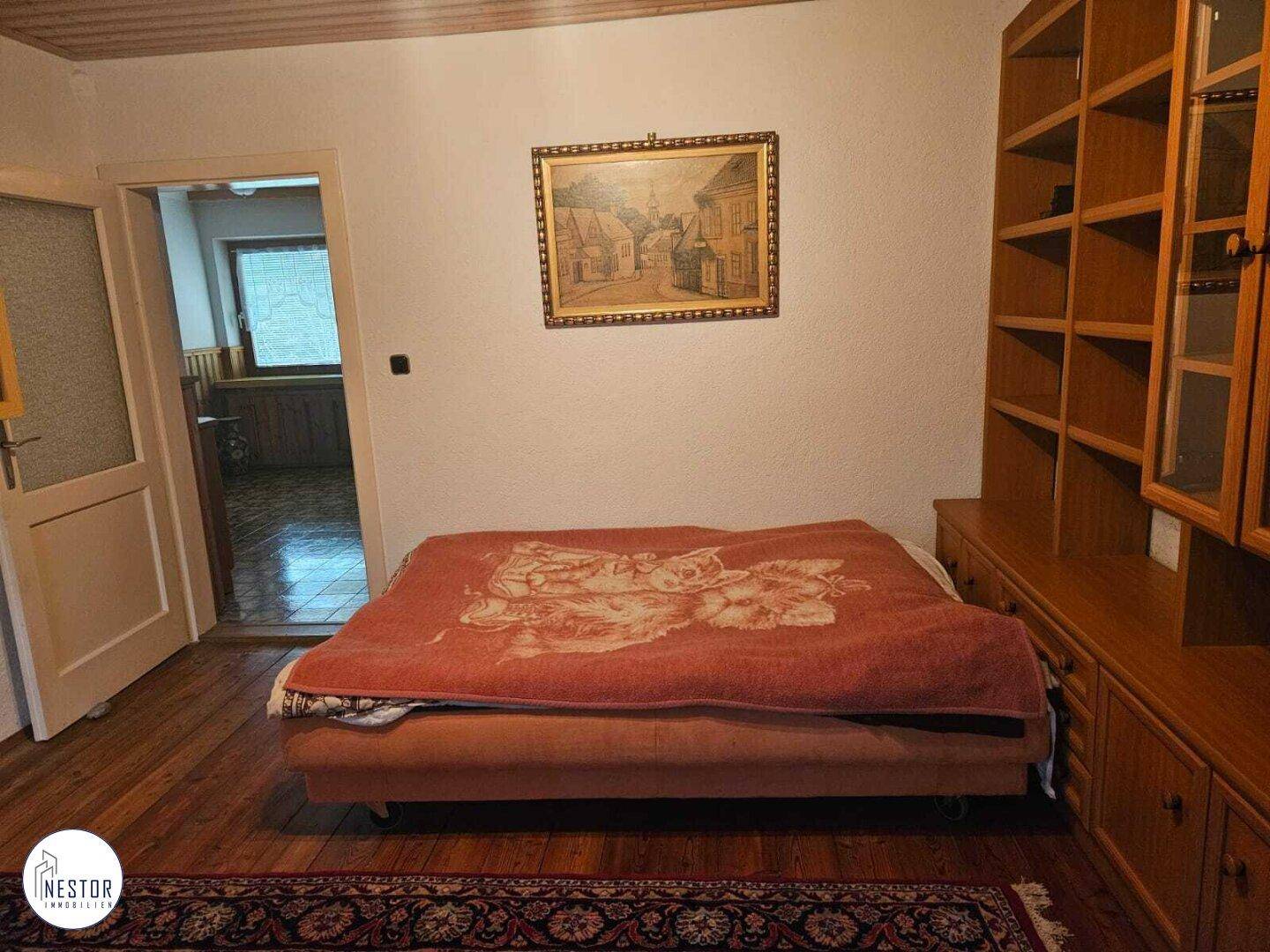 Schlafzimmer mit Holzboden, großem Bett, Holzregal und Zugang zu einem weiteren Raum.