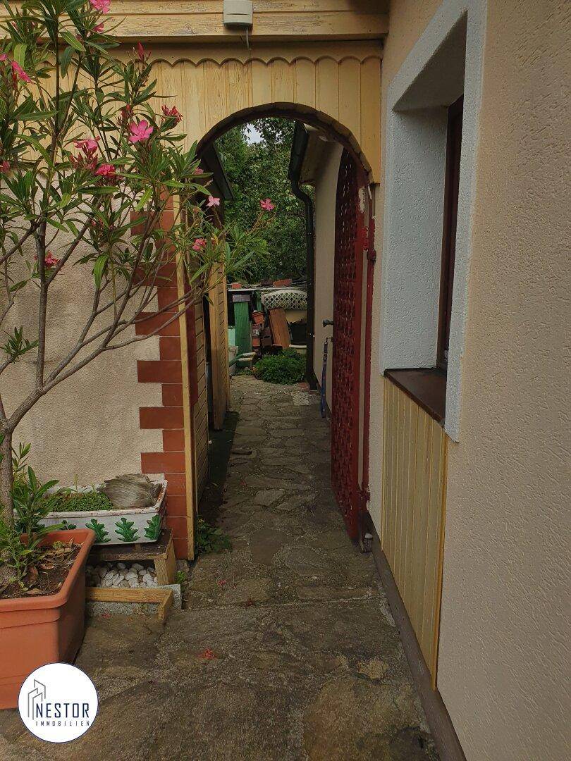 Schmaler gepflasterter Weg entlang des Hauses mit einem Torbogen und Pflanzen.