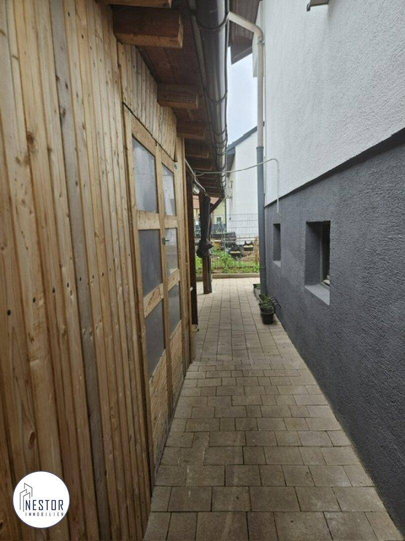 Schmaler gepflasterter Weg zwischen dem Haus und einem Holzschuppen im Außenbereich.