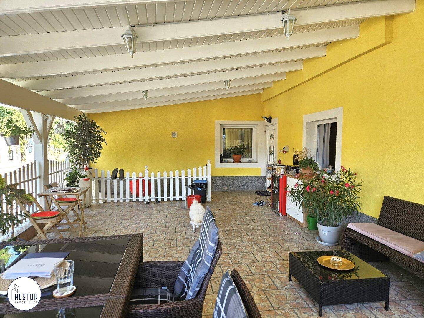 Geräumige überdachte Terrasse mit gelber Hauswand, gefliestem Boden und verschiedenen Pflanzen.