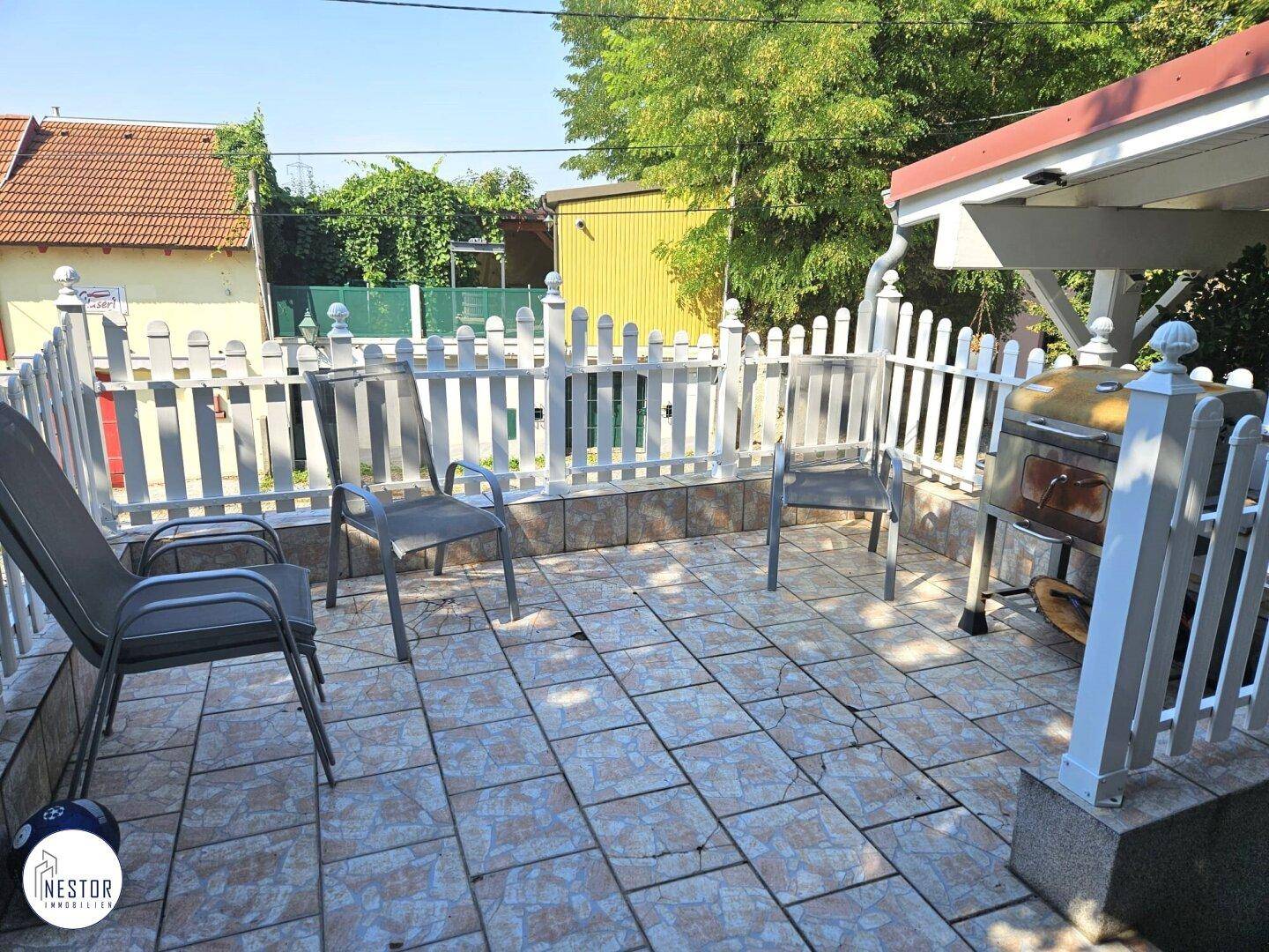 Kleiner gefliester Terrassenbereich mit Grill und Gartenstühlen, umgeben von einem weißen Zaun.