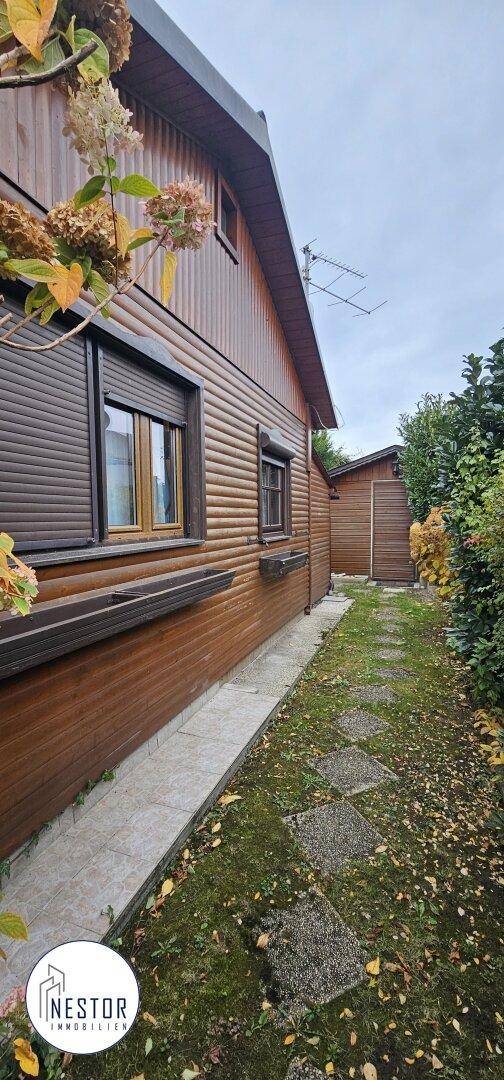 Seitenansicht des Holzhauses mit brauner Fassade, Rollläden und einem gepflasterten Weg im Garten.
