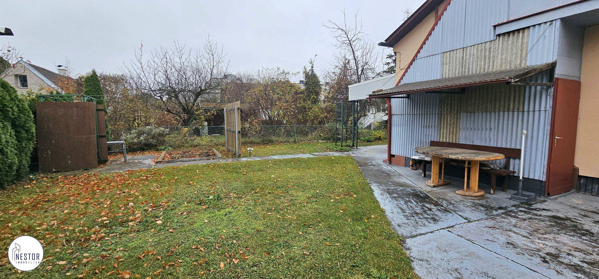 Gartenbereich mit Rasen, Bäumen und einem überdachten Sitzbereich am Haus.