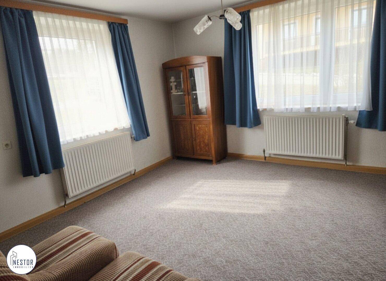 Helles Wohnzimmer mit Teppichboden, Holzschrank und blauen Vorhängen.