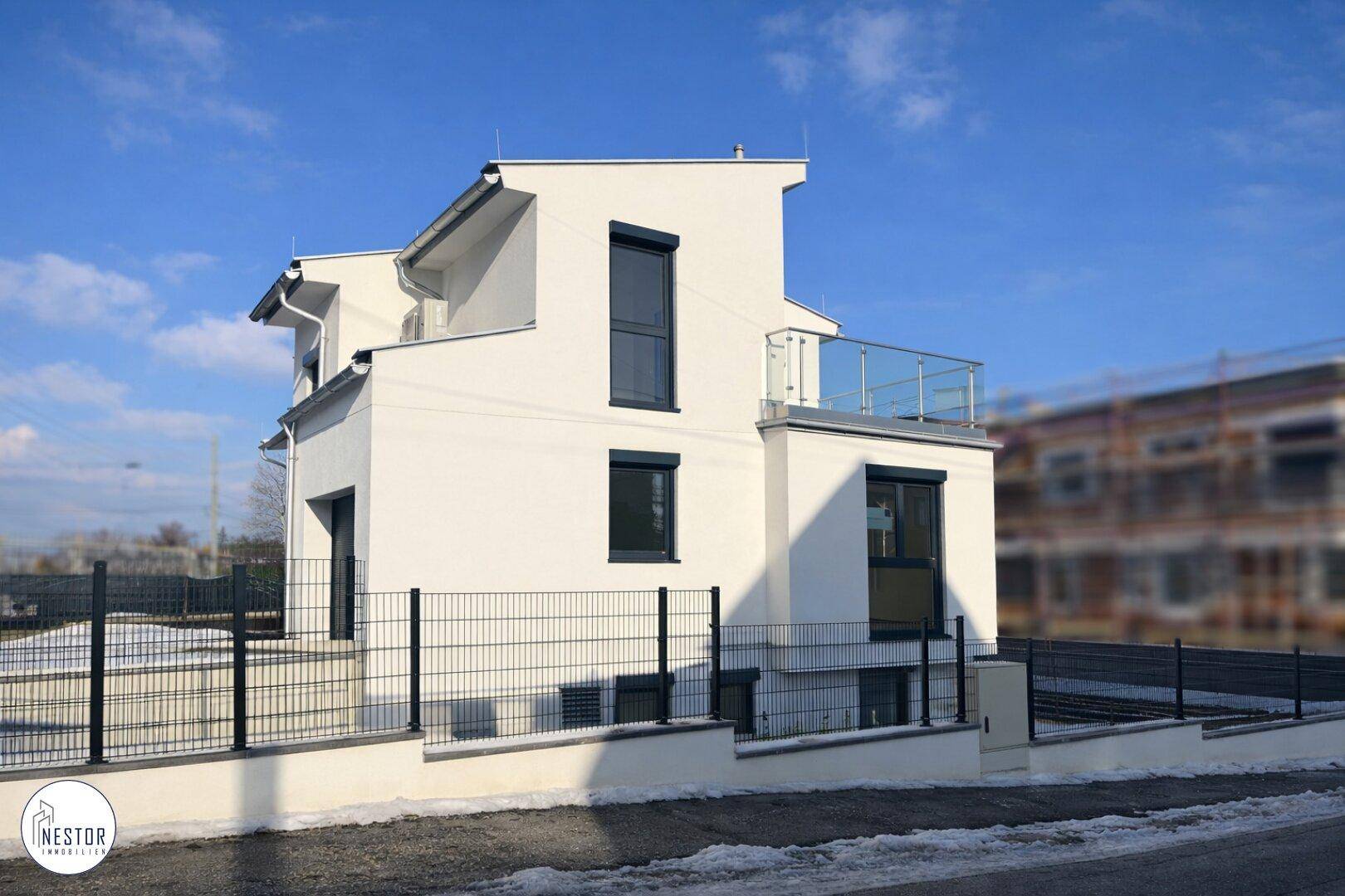 Modernes Einfamilienhaus mit weißer Fassade, dunklen Fenstern und Balkon mit Glasgeländer.