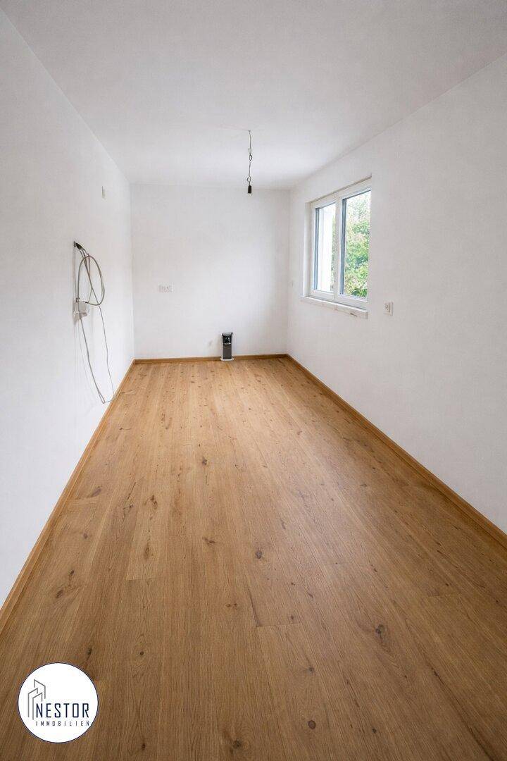 Leeres Zimmer mit hellem Holzboden und einem Fenster, das viel Tageslicht hereinlässt.