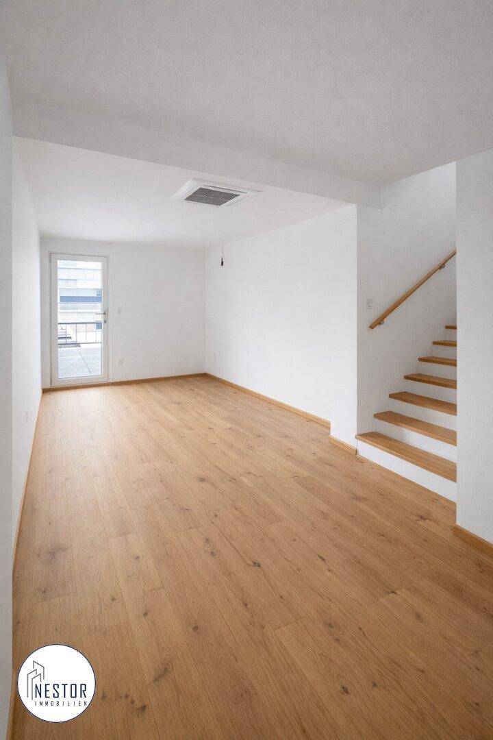 Geräumiger, leerer Raum mit Holzboden und einer Holztreppe, die in ein Obergeschoss führt.