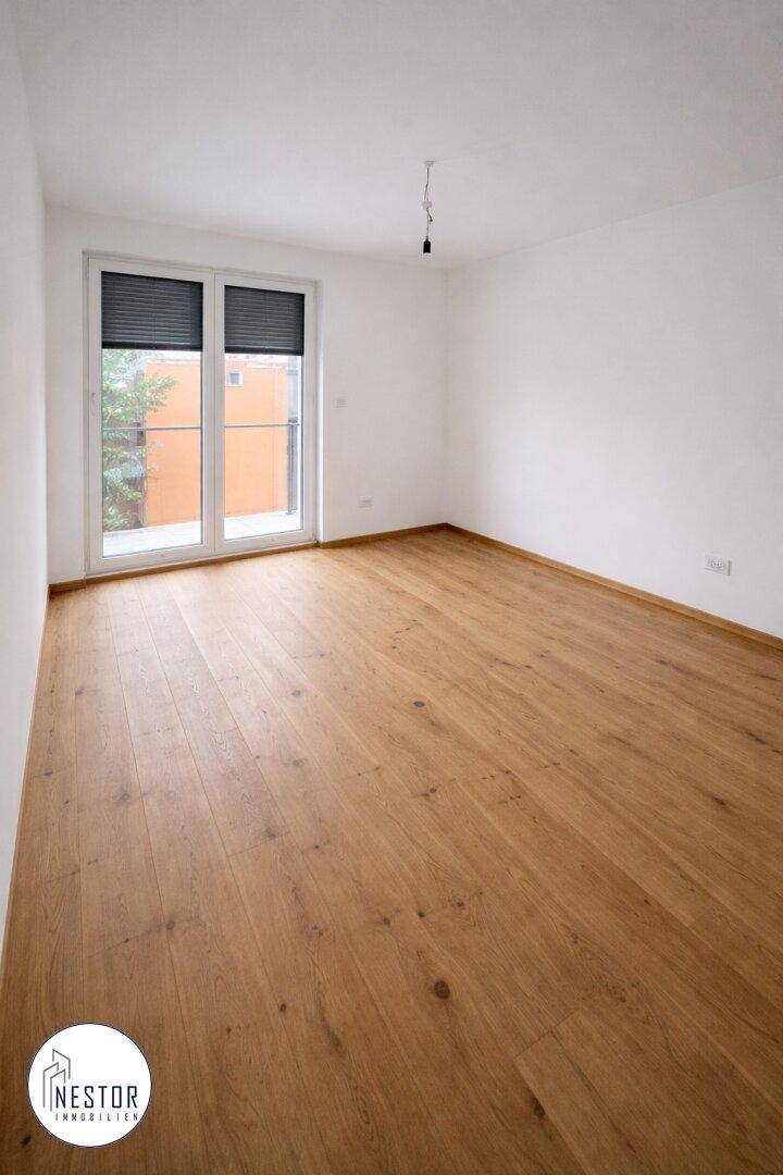 Heller, leerer Raum mit Holzboden und großer Glas-Schiebetür, die Zugang nach draußen bietet.