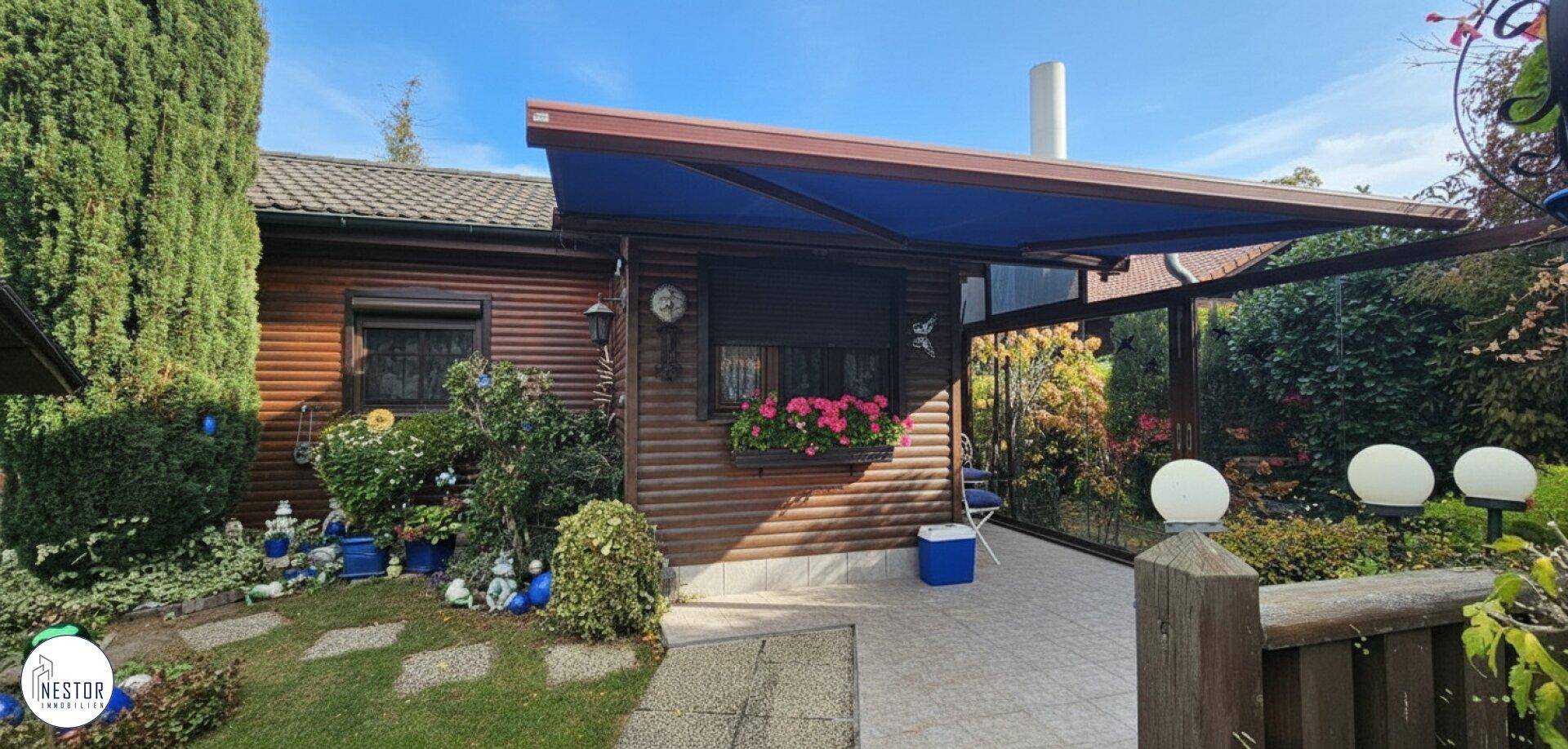 Holzhausfassade mit blauer Markise über einer gepflasterten Terrasse und Blumenkästen an den Fenstern.