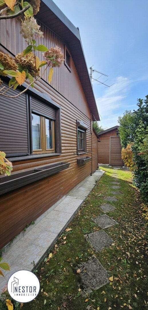 Seitenansicht des Holzhauses mit brauner Fassade, Rollläden und einem gepflasterten Weg im Garten.