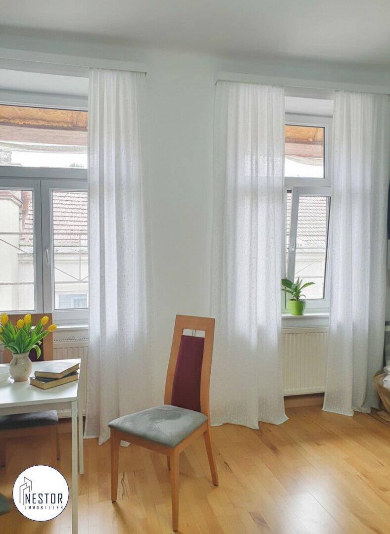 Heller Raum mit Holzboden, weißen Vorhängen und Blick auf die umliegenden Gebäude.