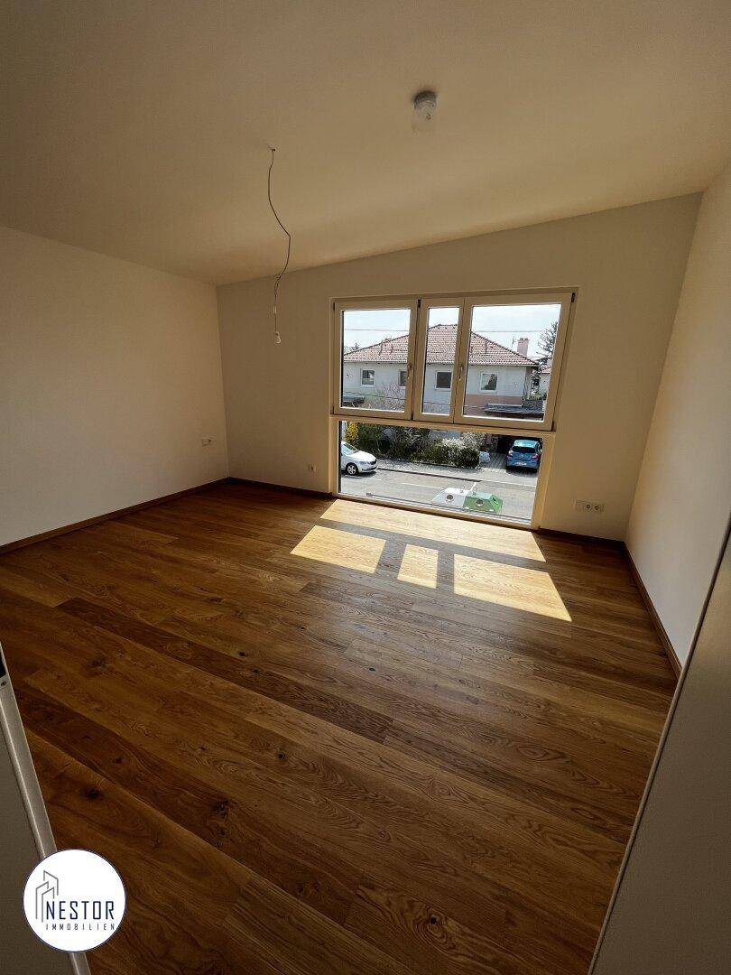 Leeres Schlafzimmer mit Holzboden und großem Fenster, das viel Tageslicht hereinlässt.