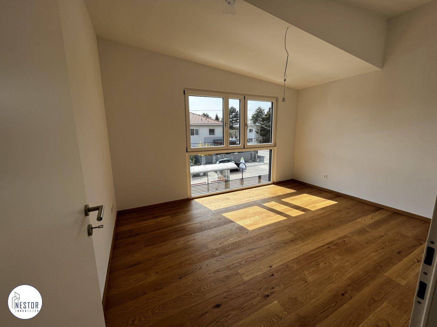 Unmöbliertes Schlafzimmer mit Holzboden und großem Fenster, das den Raum hell erscheinen lässt.