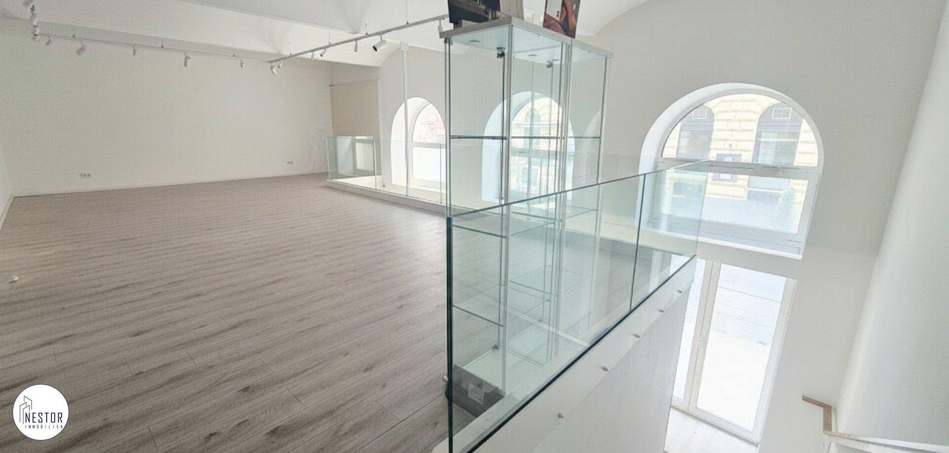 Offener Bereich mit Glasgeländer, gewölbter Decke und großen Rundbogenfenstern, die Licht hereinlassen.
