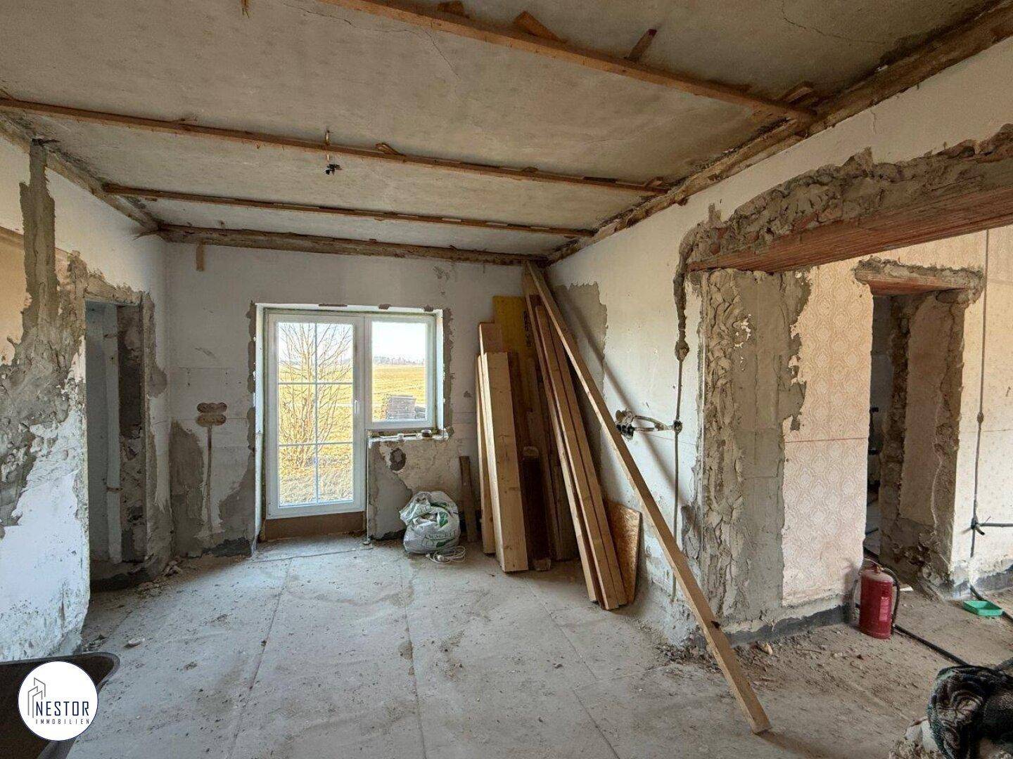 Innenraum im Rohbauzustand mit sichtbaren Deckenbalken und einem Fenster mit Blick ins Grüne.
