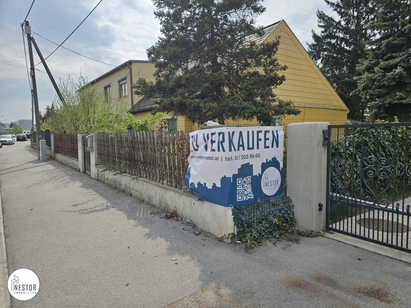 Außenansicht eines gelben Hauses mit Zaun, Tor und einem 'Zu verkaufen'-Banner an der Straße.