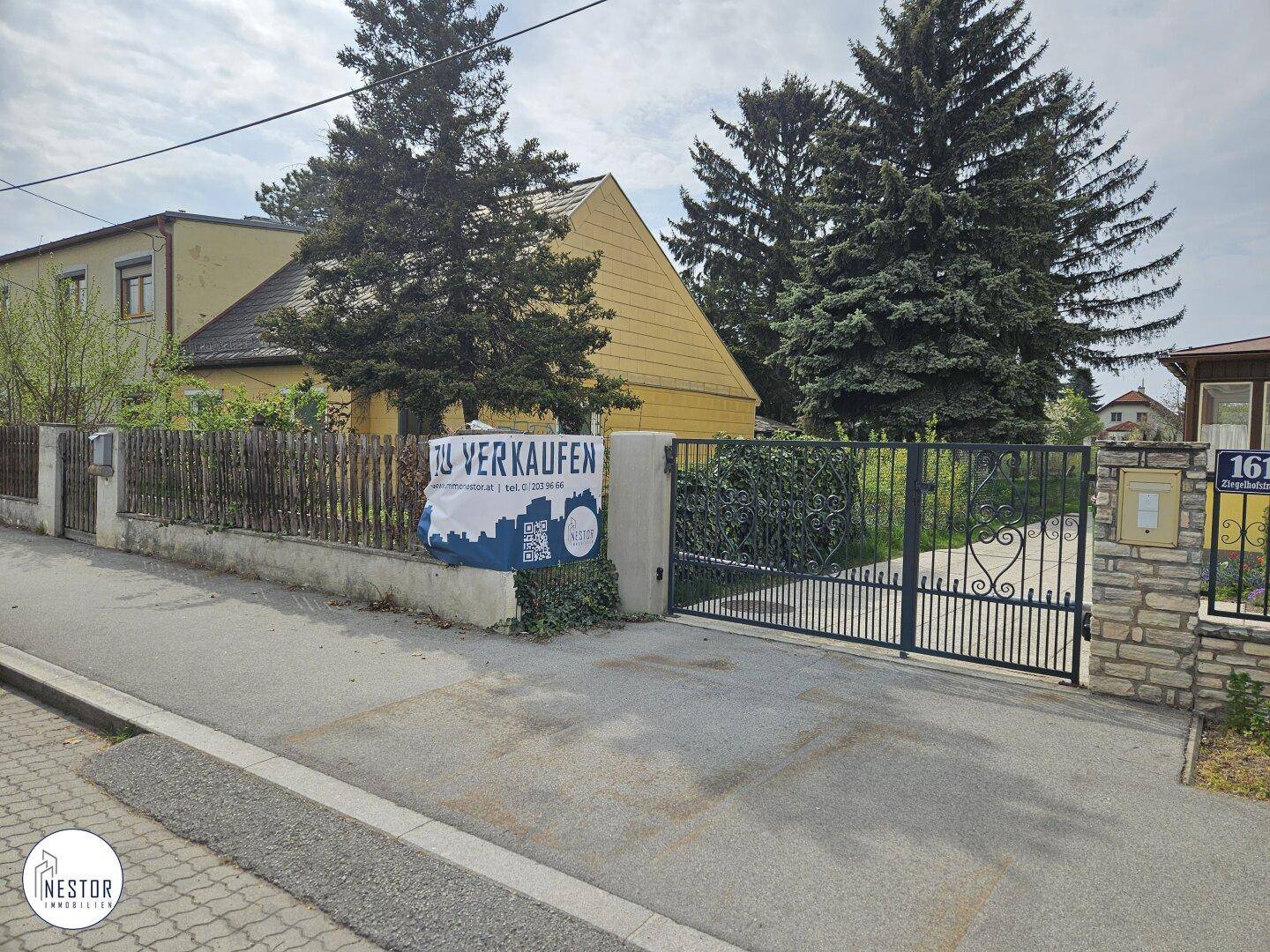 Blick von der Straße auf ein älteres gelbes Haus mit einem 'Zu verkaufen'-Schild und grüner Bepflanzung.