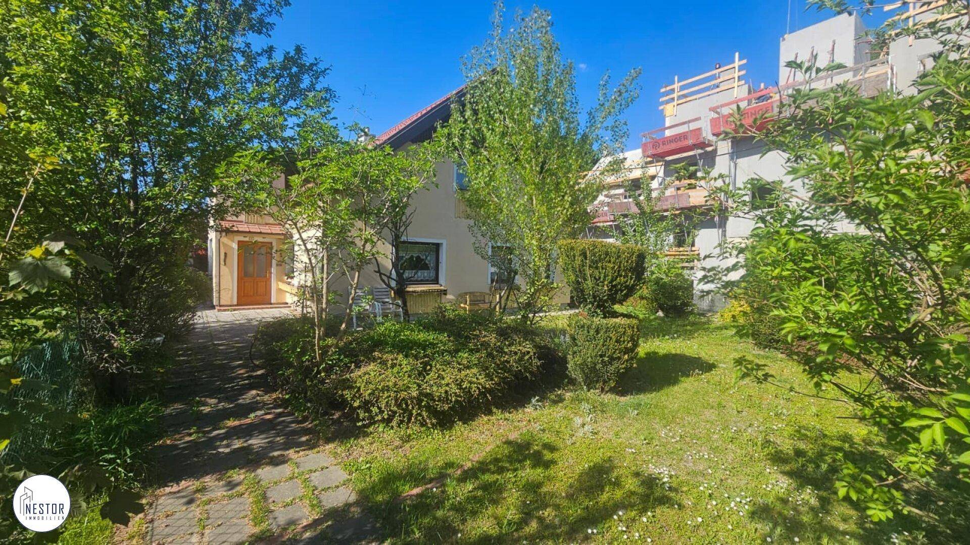Gepflegter Gartenbereich mit Rasenfläche, Büschen und einem Wohnhaus im Hintergrund unter blauem Himmel.