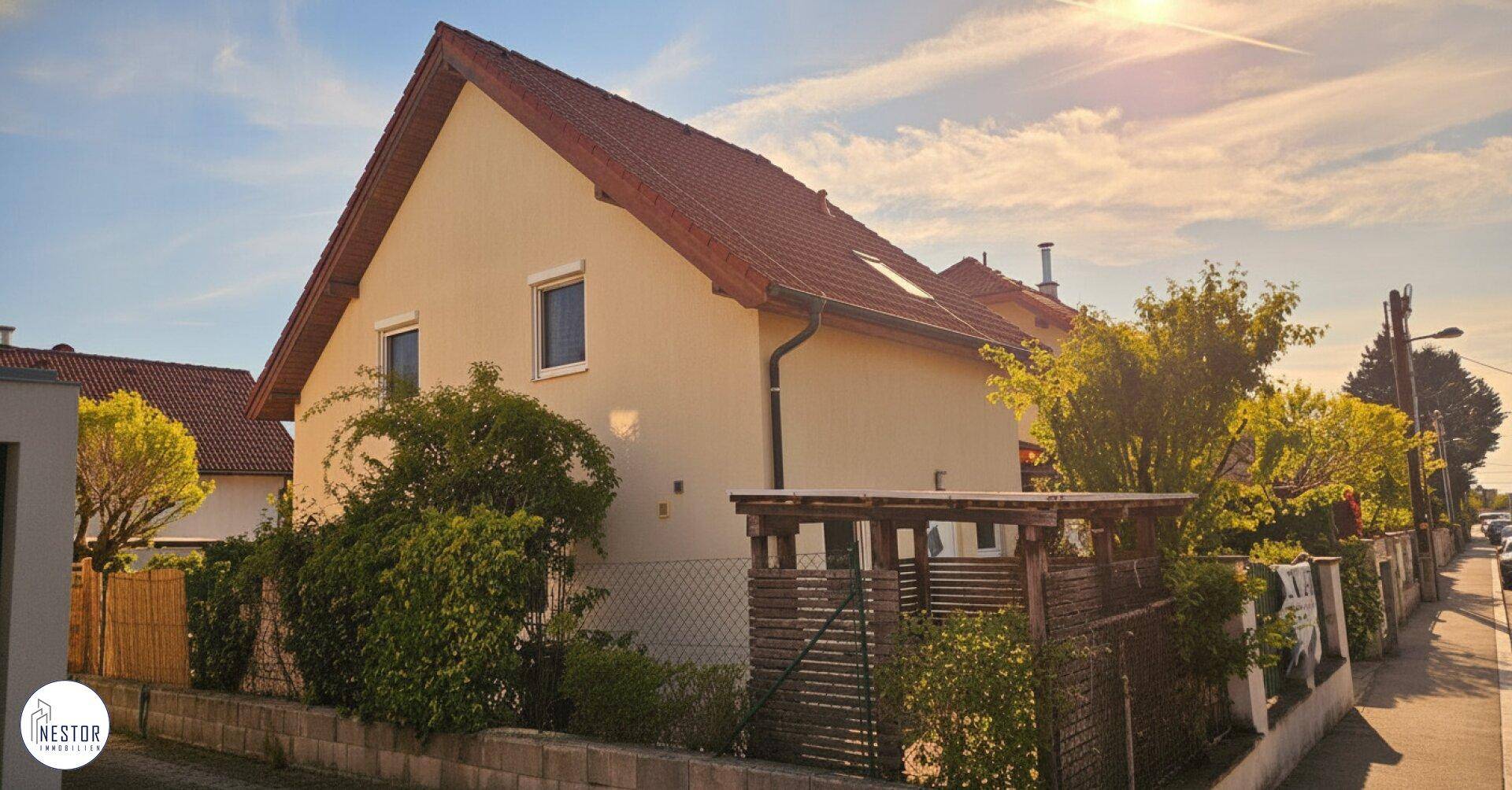 Ansicht des Hauses von der Straße bei Sonnenuntergang, umgeben von Bäumen und Hecken.