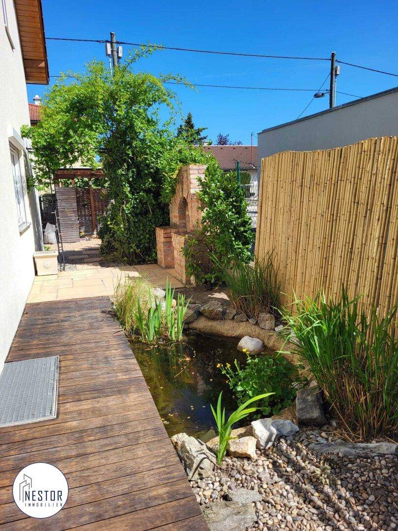Holzsteg entlang eines Gartenteichs mit Grillplatz und Bambuszaun im Hintergrund.